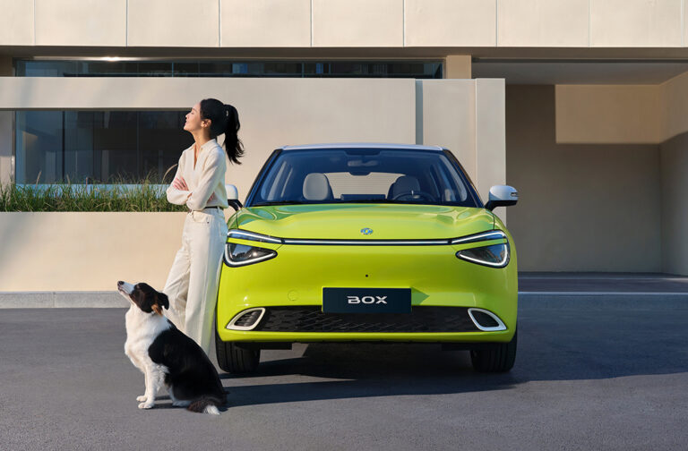 Sieviete un suns blakus spilgti dzeltenam elektroauto Dongfeng Box — mūsdienīgam pilsētas auto ar izteiksmīgu dizainu