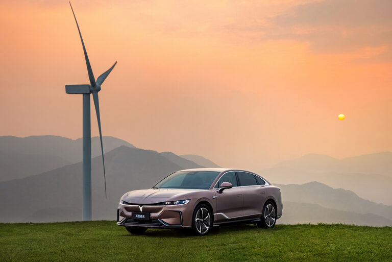 Elektroauto uz kalna pie vēja turbīnas saulrietā